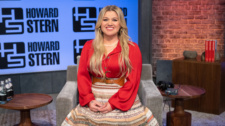 Kelly Clarkson posing for a portrait sitting at the Howard Stern Show studios in a red top and patterned skirt