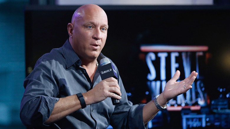 Steve Wilkos talking into a microphone with one hand extended as a monitor in the back broadcasts his name