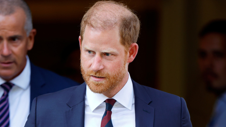 Prince Harry looking serious in a navy suit with a white shirt and a red and navy patterned tie