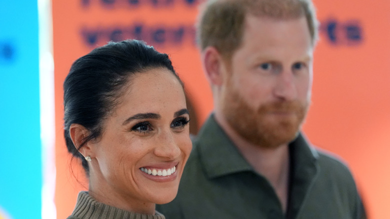 Meghan Markle smiling at someone off camera while Prince Harry watches out of focus in the background