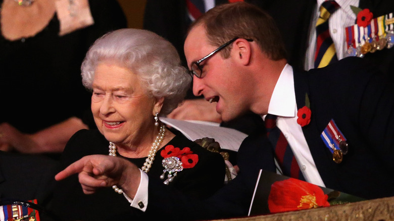 Prince William with glasses on leans over to talk to Queen Elizabeth II as he points.