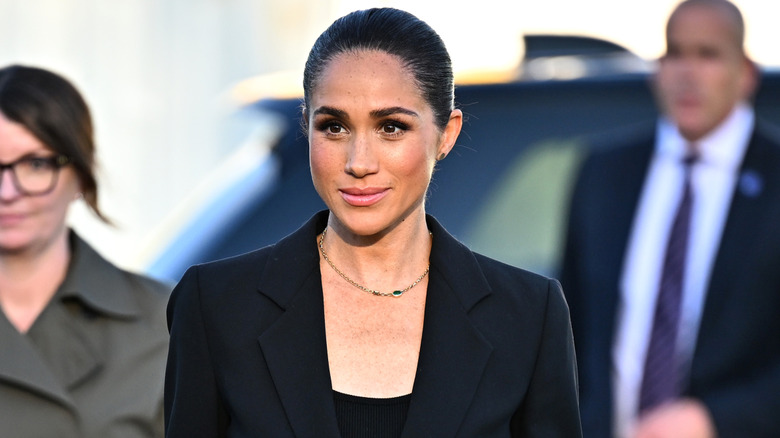 Meghan Markle smiling while looking very serious and businesslike in a black suit