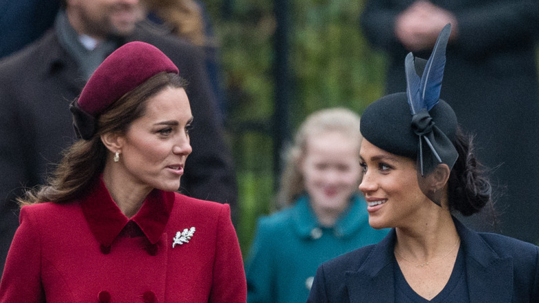 Meghan Markle wearing a navy blue coat and hat next to Kate Middleton in a dark red coat