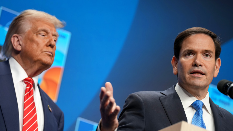 Donald Trump wearing a suit and red tie looking at Marco Rubio, who's in a suit and blue tie, while he speaks with the press.