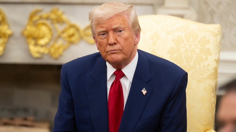 Donald Trump sitting in the Oval Office in a navy suit with a red tie
