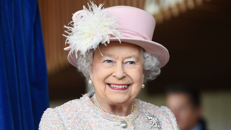 The late Queen Elizabeth II attends an event wearing a tweed jacket and a pink hair with a feather