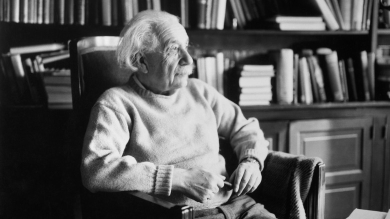 A black-and-white photo shows Albert Einstein looking on while sitting in a library