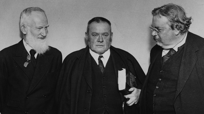 Hilaire Belloc looking serious and standing between George Bernard Shaw and G.K. Chesterton who are smiling at him