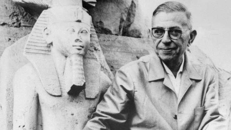 Jean-Paul Sartre smiling and sitting next to an ancient Egyptian statue