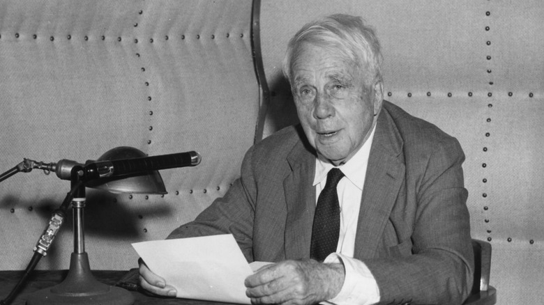 Robert Frost sitting in front of a microphone in a suit with a sheet of paper in his hands