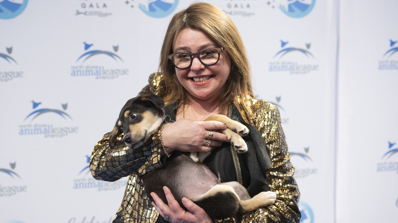 Rachael Ray smiling while holding a dog and wearing glasses and a gold jacket