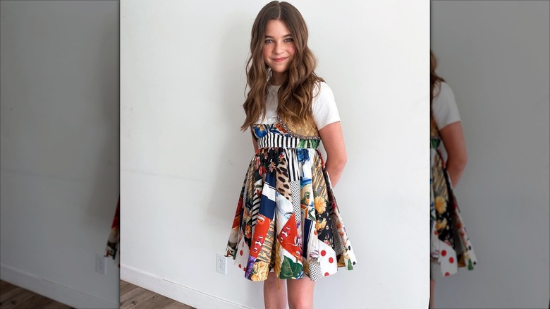 Raegan Revord in a multicolored dress smiles in front of a white wall.