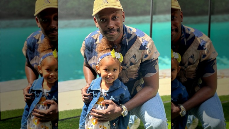 Lamorne Morris smiles in a yellow hat holding his smiling daughter in a dress in front of him outside
