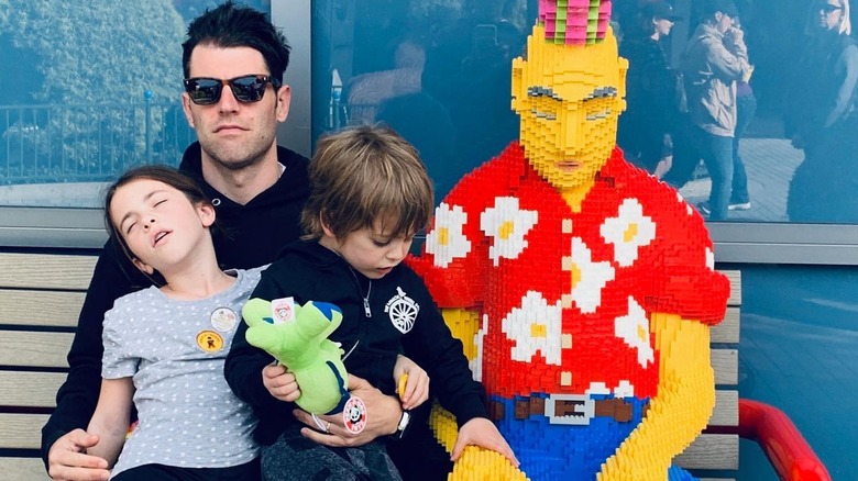 Max Greenfield holds his son and daughter who look tired on a bench at Legoland next to a Lego statue