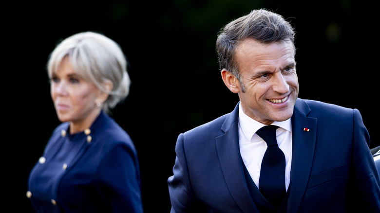 Emmanuel Marcon smiling in a suit with Brigitte Macron in the background