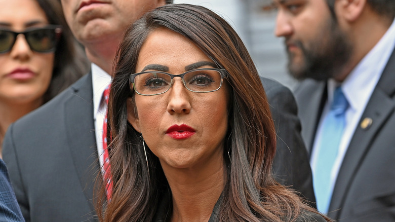 Lauren Boebert staring while wearing glasses, a purple shirt and black blazer.