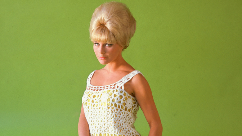 Elke Sommer, German actress, wearing a crochet minidress with a beehive hairstyle and sitting on the floor in a studio portrait, against a green background, circa 1965.