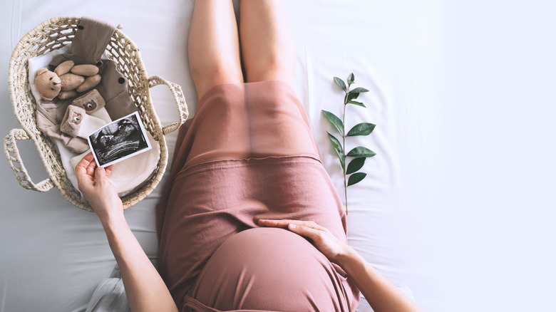 woman sitting on the ground in between a basket and a branch of leaves holding onto her pregnant stomach and an ultrasound