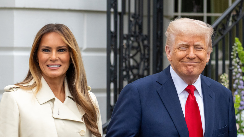 Donald Trump smiling in a suit and red tie while holding the hand of Melania Trump, who's wearing a tan trench coat.