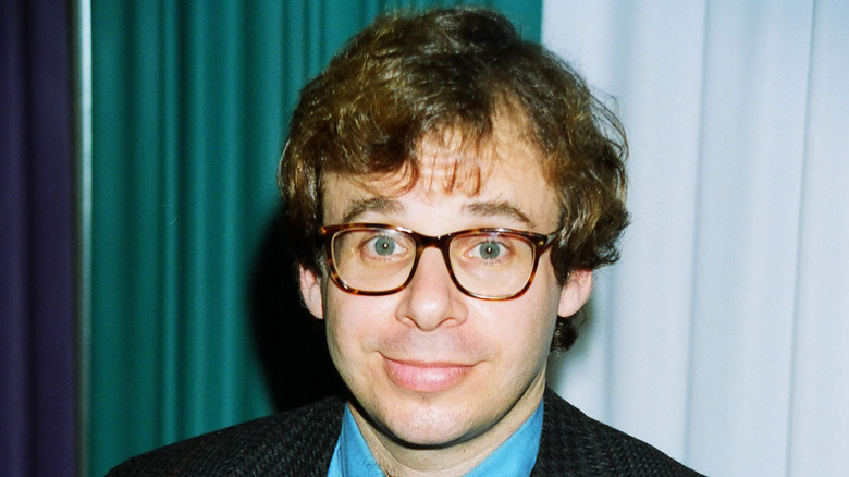 Rick Moranis smiling while posing for a photo in a light blue shirt and glasses
