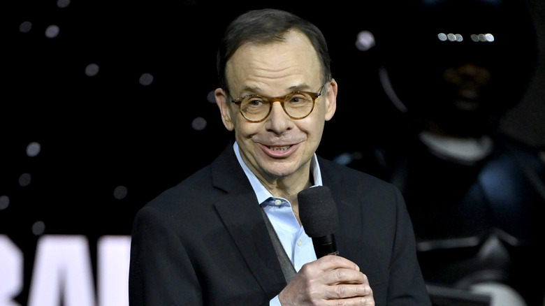 Rick Moranis speaking onstage at CinemaCon in a black suit with a light-blue shirt