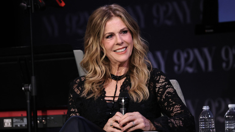Rita Wilson smiling in a black lacy top with choker necklace while holding a microphone
