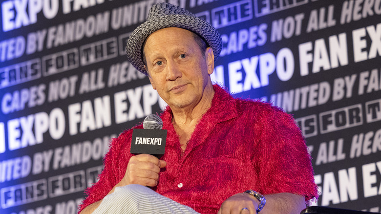 Rob Schneider holding microphone at event while wearing a hat and a red furry shirt