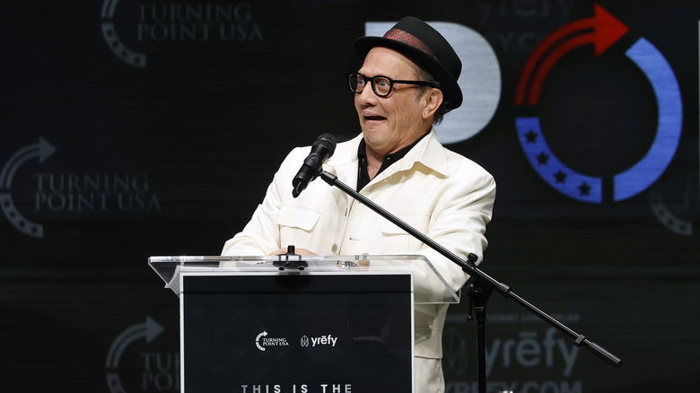 Rob Schneider looking shocked in a white suit in front of a podium at Turning Point USA event