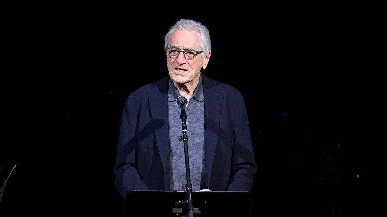 Robert De Niro speaking from a podium in a blue blazer and glasses