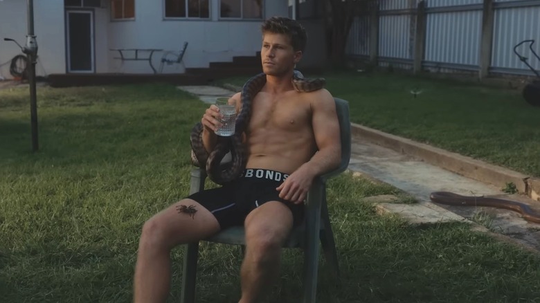Robert Irwin holding a water glass in his underwear with a snake draped around his neck and a tarantula on his thigh