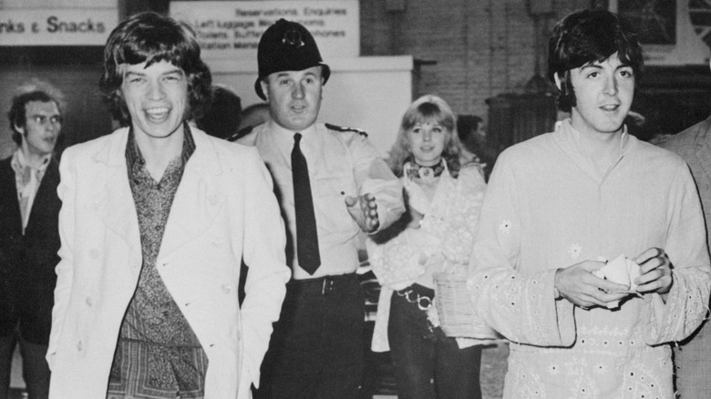 A black-and-white photo of Mick Jagger and Paul McCartney walking alongside one another with others in the background