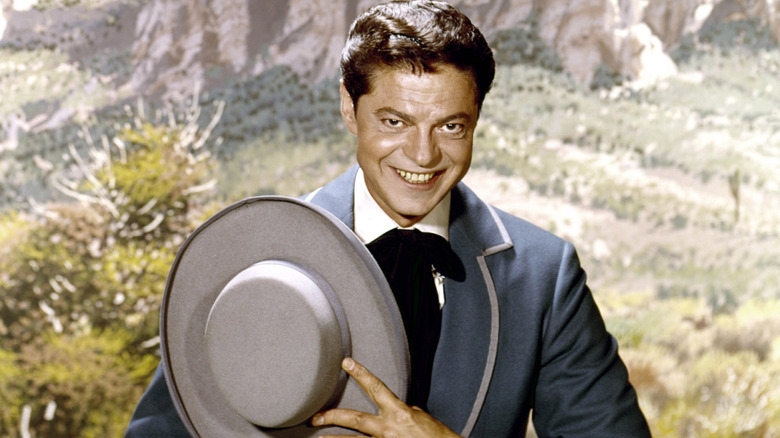 Ross Martin smiling and holding a wide-brimmed hat in a posed studio portrait