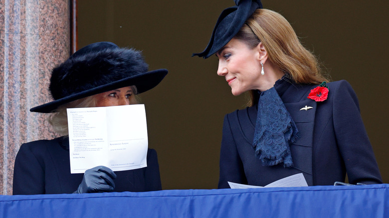 Queen Camilla covering her mouth with paper while speaking to Kate Middleton