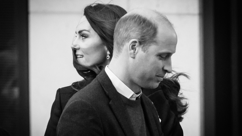 A black-and-white photo of Prince WIlliam looking to the left while Kate Middleton stands behind him and looks to the right
