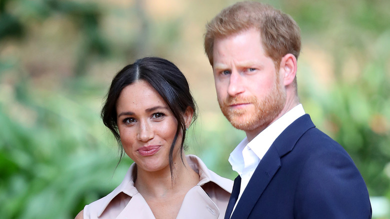 A smiling Meghan Markle standing next to a serious-looking Prince Harry