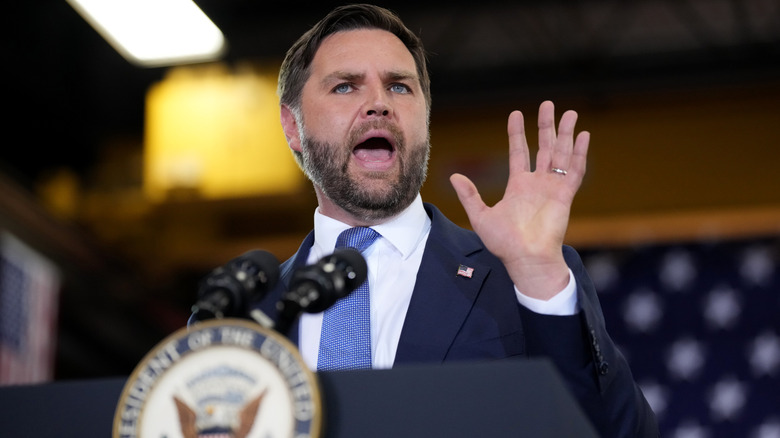 JD Vance raising his hand while delivering a speech in a navy suit with a blue tie