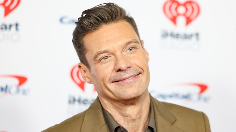 Ryan Seacrest in a tan suit, smiling with his head cocked to the side.