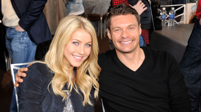Julianne Hough in a white chair, leaning into the arms of Ryan Seacrest in a black sweater.
