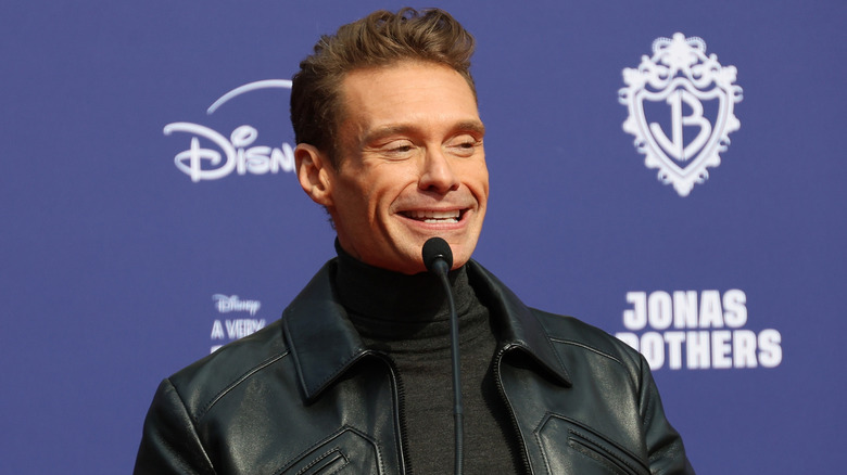 Ryan Seacrest wearing a turtleneck and black leather jacket while speaking into a microphone
