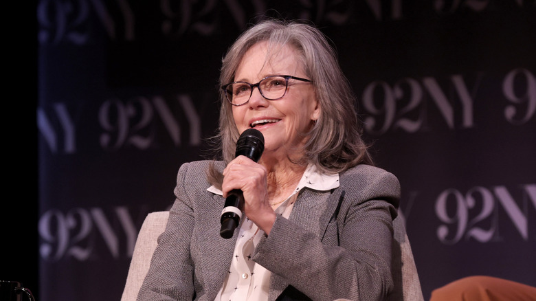 Sally field in a grey suit and star studded white blouse holding a microphone while addressing a crowd