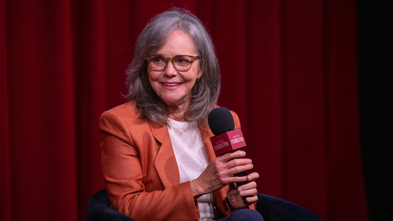 Sally Field holding a microphone wearing an orange blazer with a white shirt.