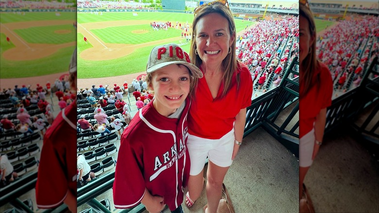 Sarah Huckabee Sanders posing with young son at a baseball game, the field in the background, with Sanders wearing a red top and short white shorts