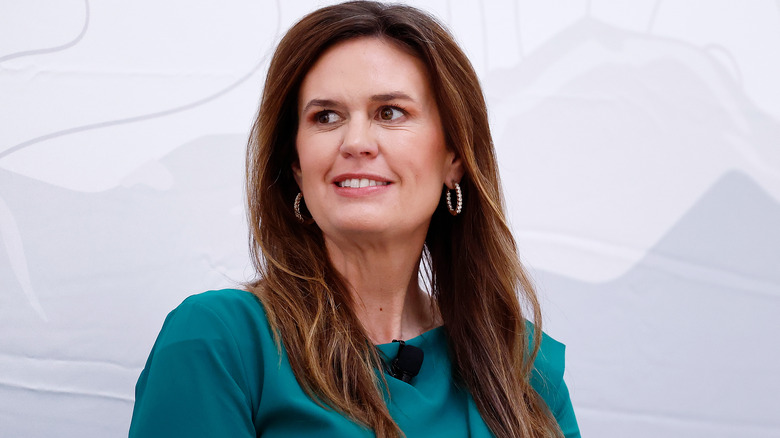 Sarah Huckabee Sanders wearing a green top, looking to the side and smiling in front of a white background