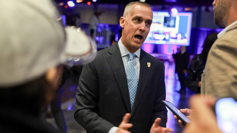Corey Lewandowski looking upset as he gestures while speaking to reporters in a dark suit