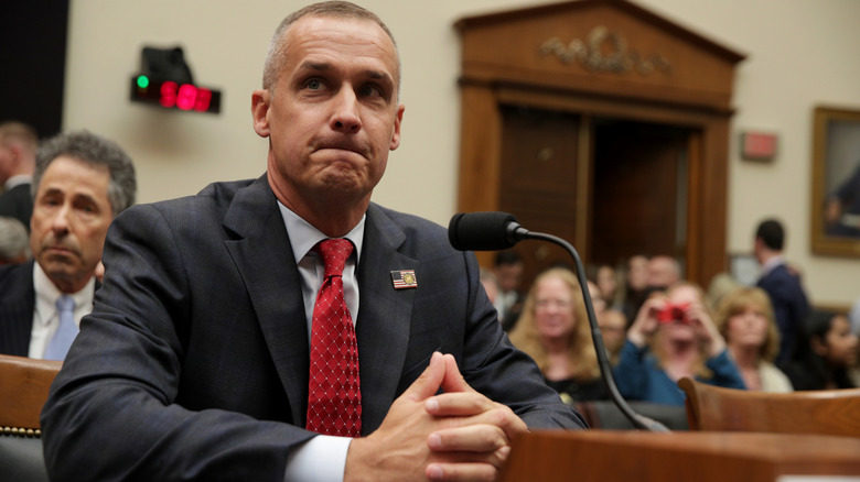 Corey Lewandowski pursing his lips and steepling his thumbs in a dark suit in front of a microphone