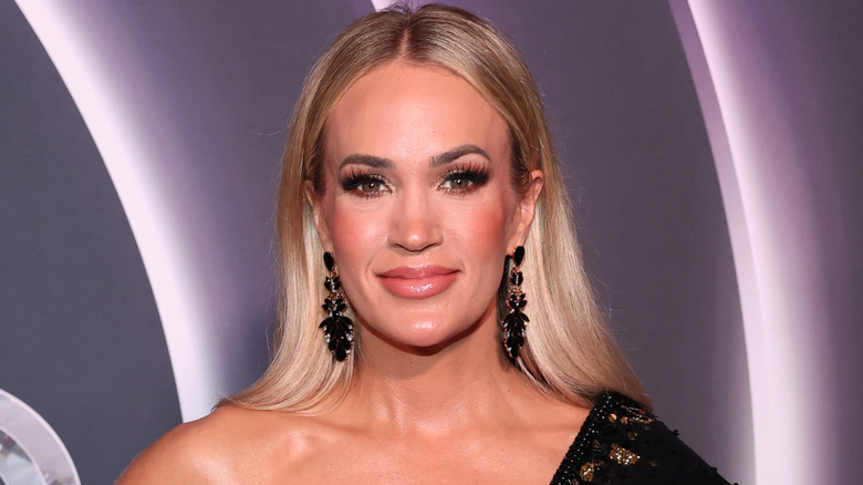 Carrie Underwood smiling while posing at an event in a black off the shoulder dress and matching dangly earrings