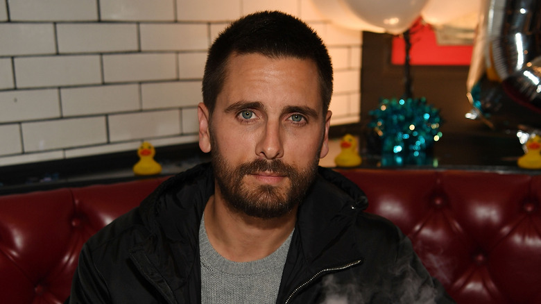 Scott Disick sitting in a red booth in a black jacket with a grey sweater underneath