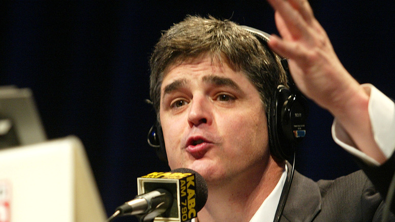 A younger Sean Hannity doing his talk show live while wearing a grey blazer, white collared shirt, and a pair of headphones.