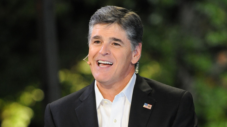 Sean Hannity smiling while wearing a microphone earpiece against green background