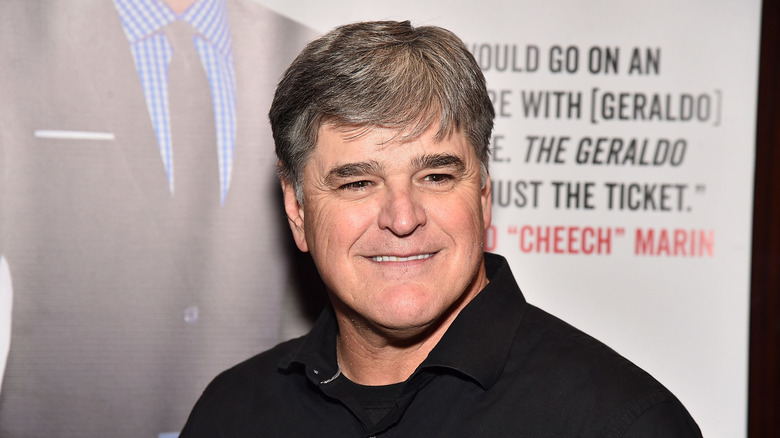 Sean Hannity in black outfit smiling in front of a backdrop with text and an image of a suit